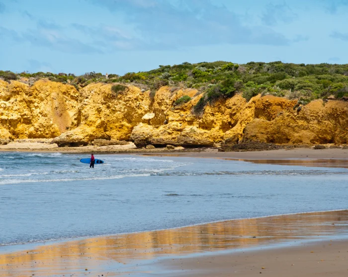 Het strand bij Torquay in Australië, met een surfer in het water en goudkleurige kliffen langs de kust