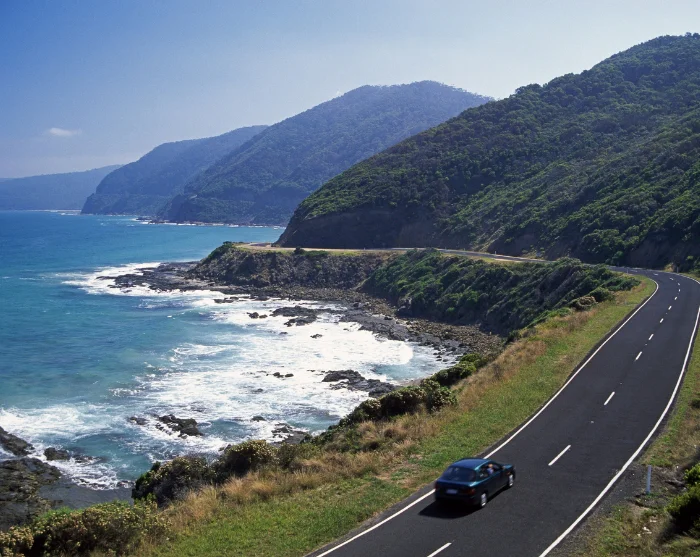 De Great Ocean Road in Australië, een kronkelende kustweg waar een auto tussen groene heuvels en de blauwe zee rijdt