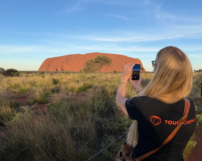 Vrouw in een TourCompass-shirt die Uluru in Australië fotografeert, omringd door het groene woestijnlandschap
