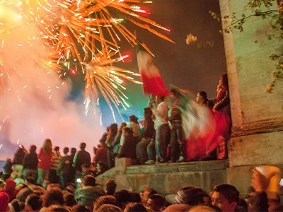 Een grote groep mensen viert de Mexicaanse Onafhankelijkheidsdag in het centrum van een stad, met vlaggen en vuurwerk